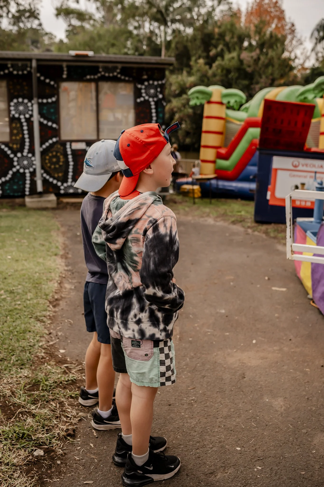 Three boys standing in line at an outdoor event, watching a colorful inflatable slide.