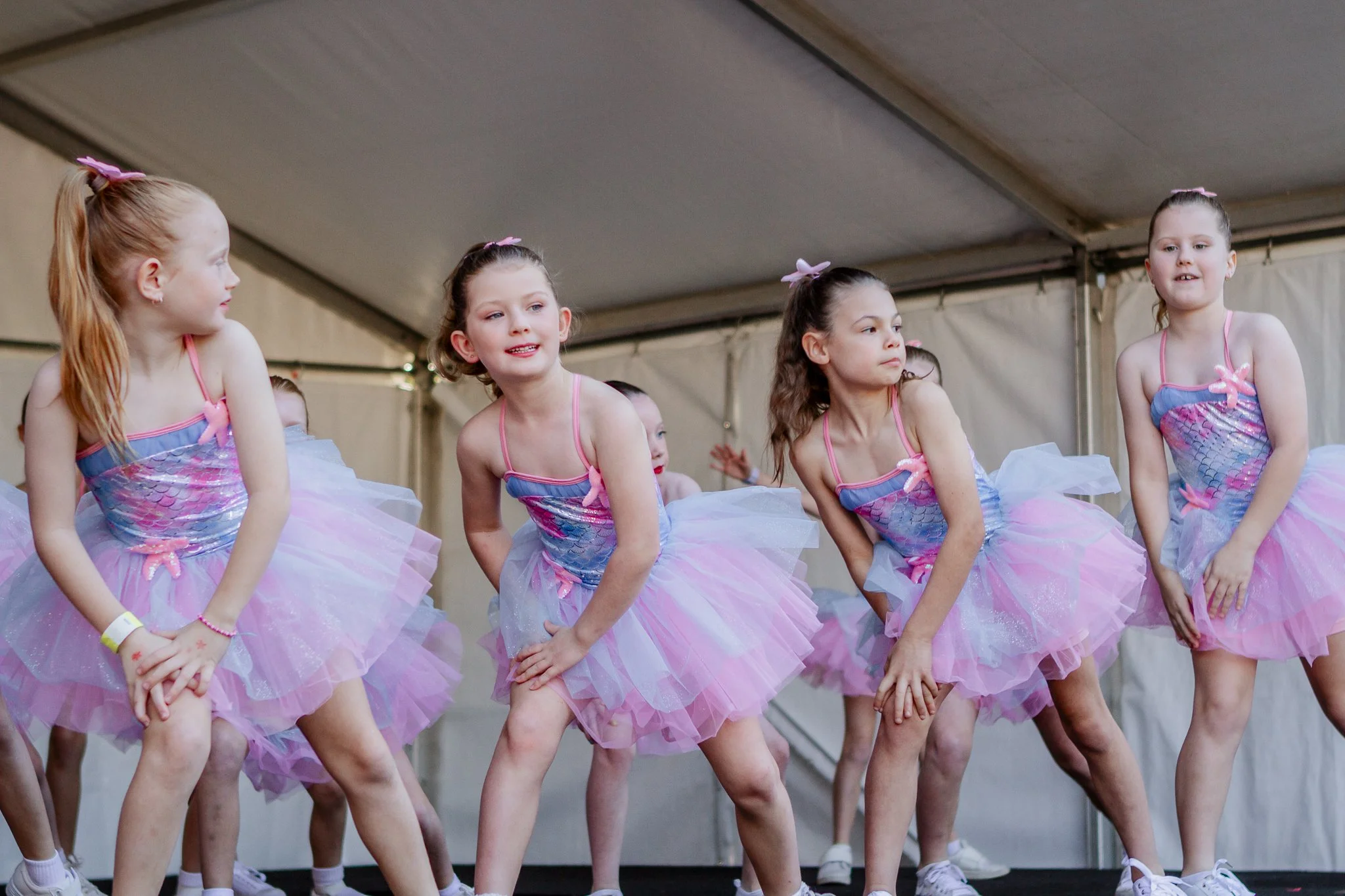 Four young girls in pink and blue tutu dresses performing a dance on stage under a tent.