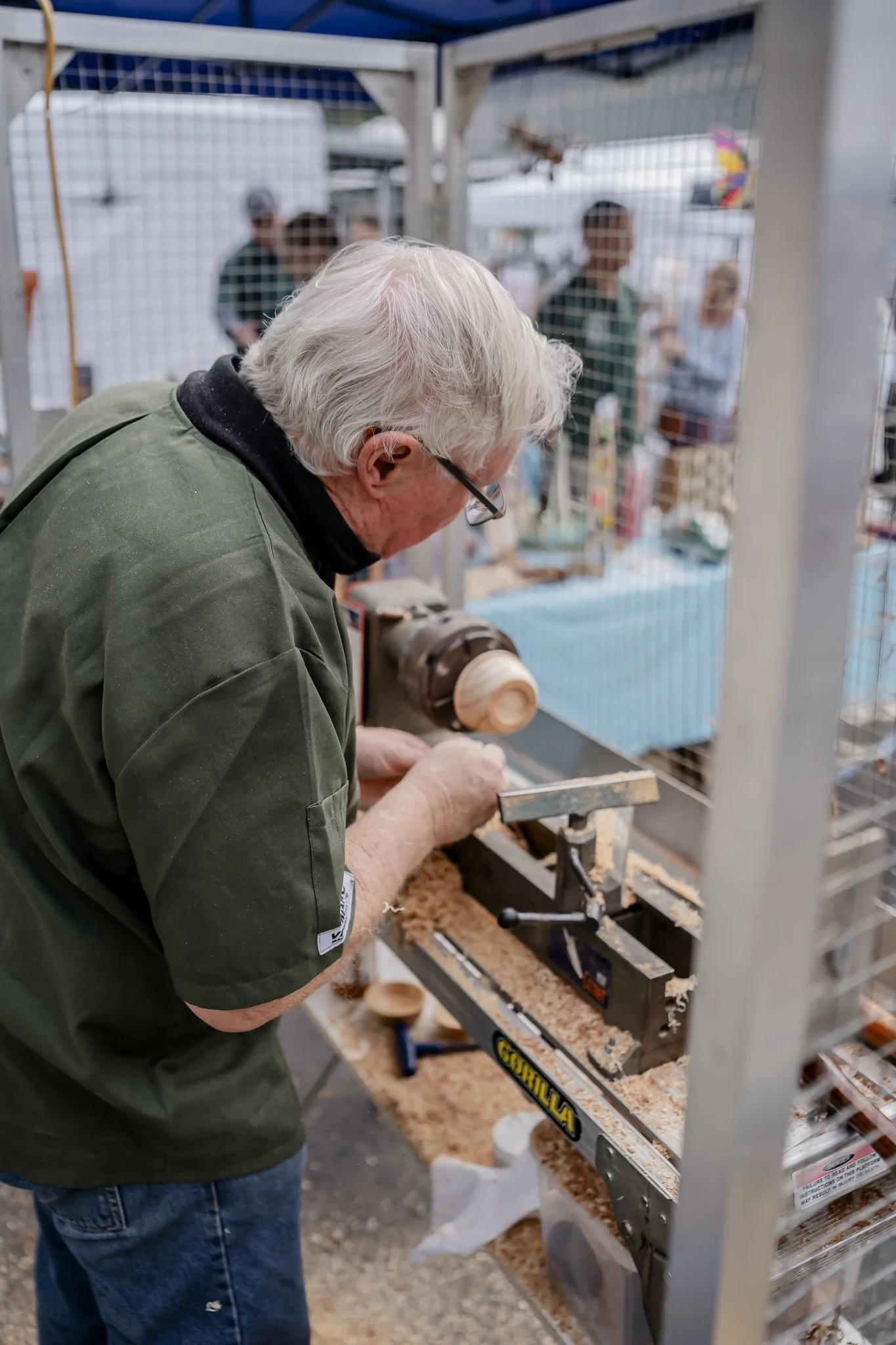 Older man working on a woodworking lathe at a craft fair, carving a small wooden object, with a wire mesh enclosure and blurred onlookers in the background.