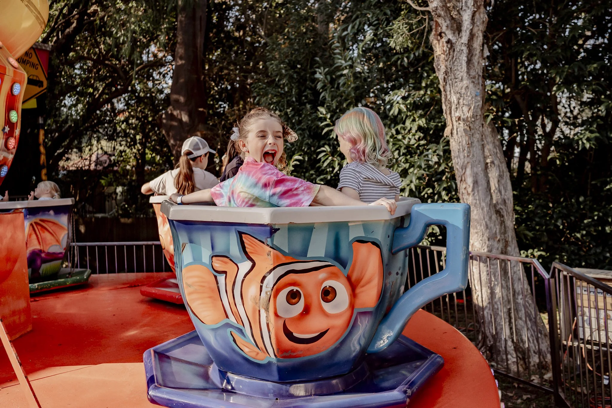 Children on a colorful amusement ride teacup with Nemo from Finding Nemo painted on it, in an outdoor setting with trees in the background.