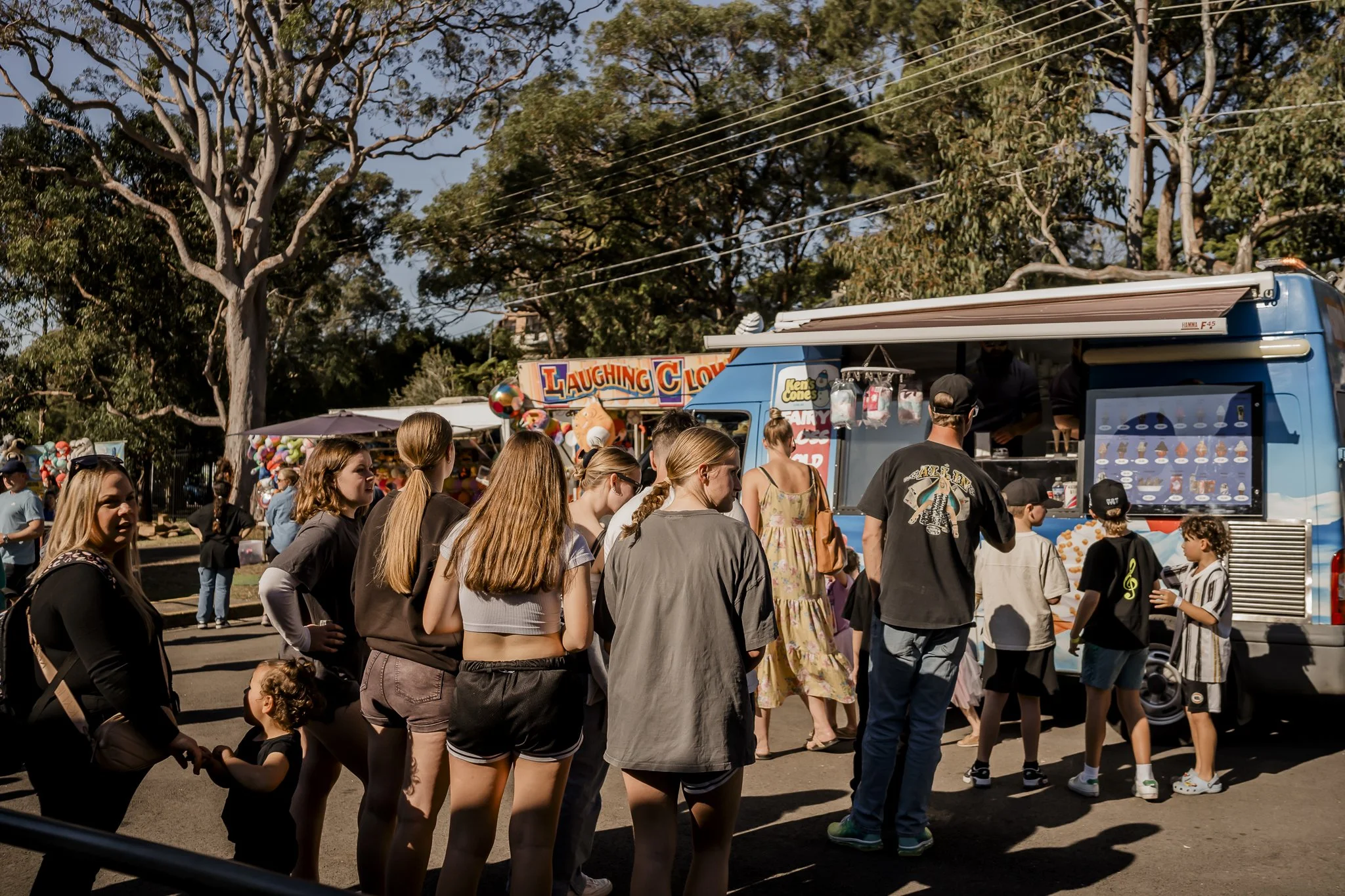 People standing in line at a food truck at an outdoor fair or festival, with trees and other vendor booths in the background on a sunny day.