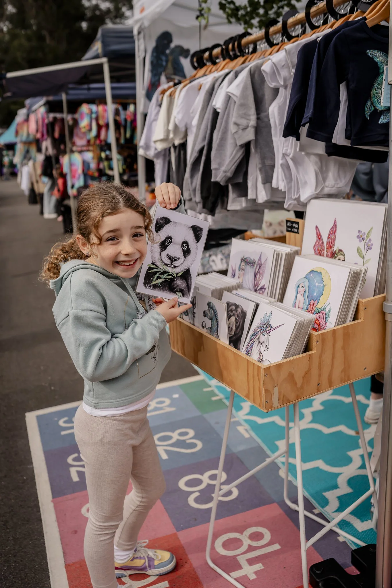 A young girl at an outdoor market stall holding a picture of a panda. The stall displays various illustrated art prints, and there are clothing items hanging on racks behind her. The girl is smiling, wearing a light green hoodie, light pink leggings,