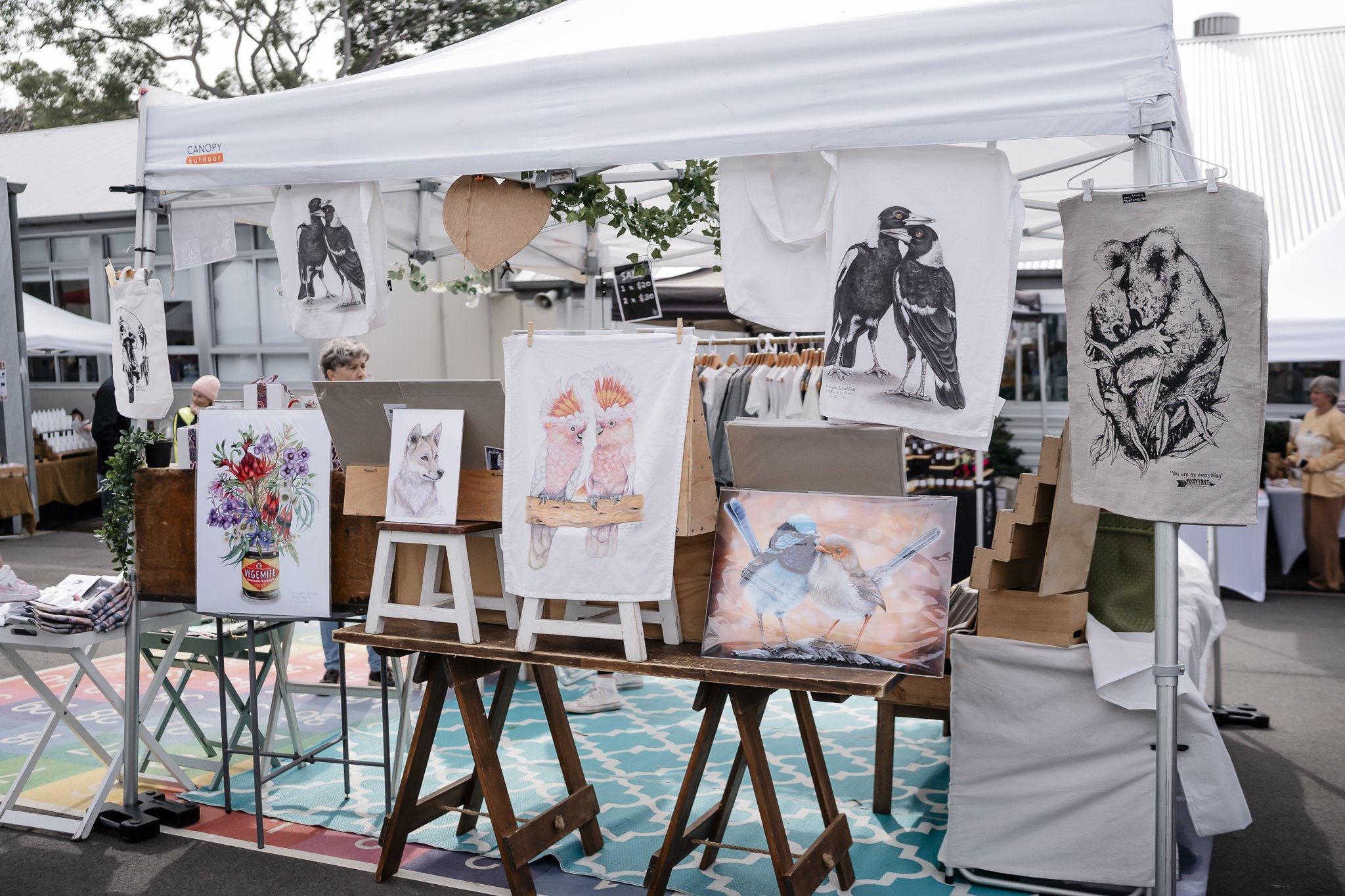 An outdoor art stall displaying various prints and illustrations of animals, including owls, wolves, bears, birds, and other wildlife, at the Oyster Bay Art and Craft Festival