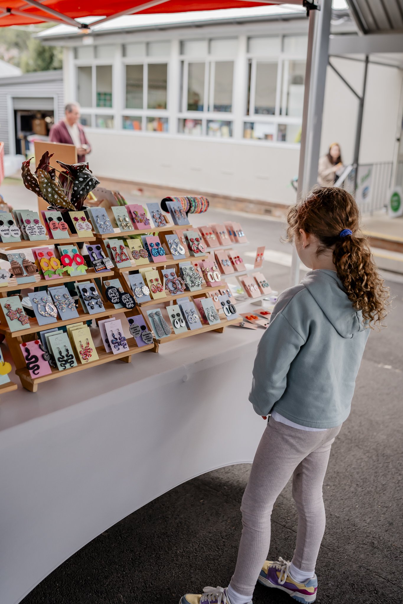 A young girl with curly hair wearing a gray hoodie and beige leggings stands in front of a jewelry display at an outdoor market. The display has earrings arranged on small cards on a tiered wooden stand. In the background, there is a man looking at p