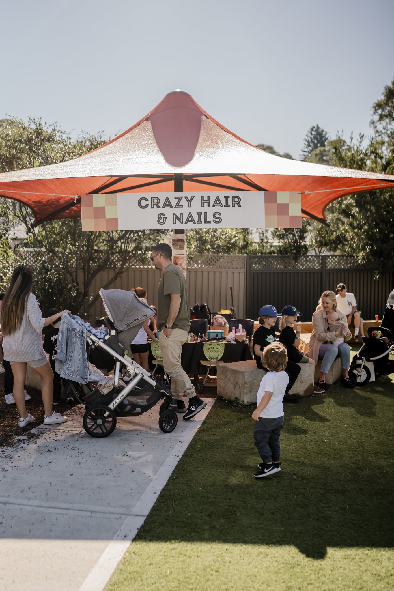 Outdoor marketplace under a large red and white canopy with a sign reading 'Crazy Hair & Nails'. People, including children and adults, are gathered, some sitting on stone benches and others standing or walking, with a stroller and various items on t