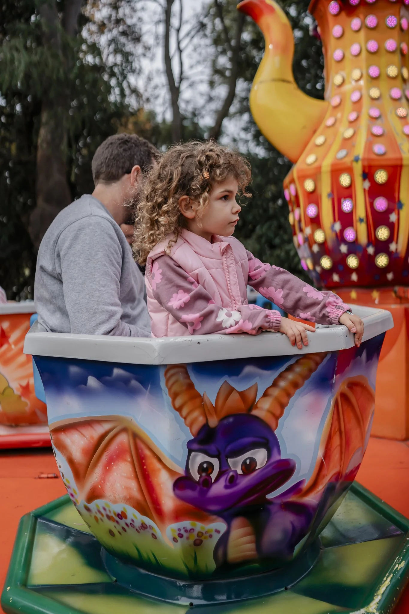 Two children, one girl with curly hair wearing a pink jacket, riding in a colorful teacup ride with dragon artwork at amusement park.