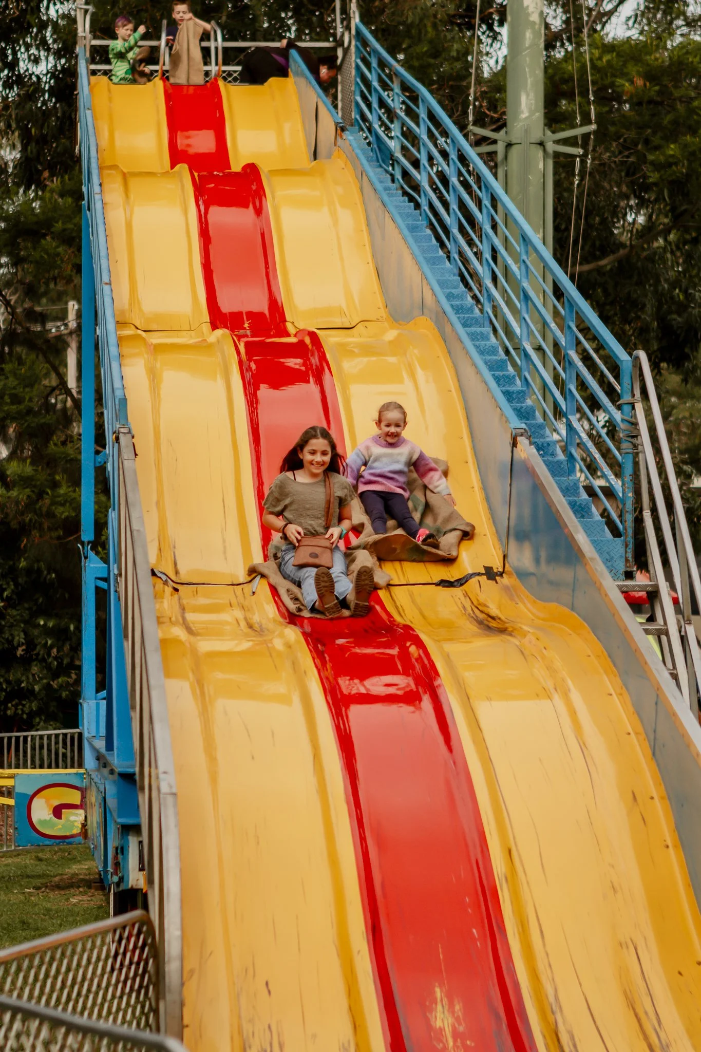Two girls riding down a yellow and red slide at an amusement park, smiling and holding on to a burlap sack; other children are at the top of the slide at the Oyster Bay Art and Craft Festival.