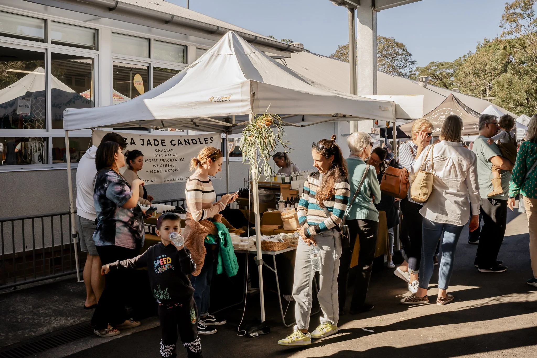 People shopping at an outdoor market stall selling candles and home fragrances, with tents and a building in the background on a sunny day at The Oyster Bay Art and Craft Festival