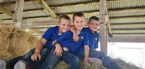 Three young boys sitting together on a hay bale inside a barn, smiling.