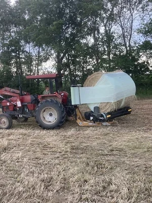 A red tractor pulling a large, white, rounded hay bale storage cover on a farm field with trees in the background.