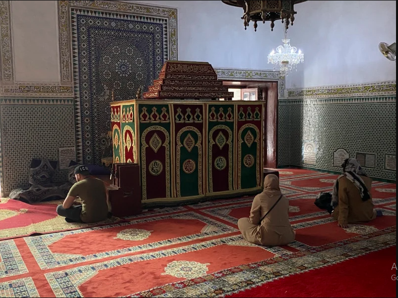 Interior of the mausoleum of Sīdī Muḥammad al-Jazūlī in Marrakesh, showing the catafalque draped in green and gold brocade, zellij-tiled walls, red carpets, and visitors seated in prayer and contemplation.