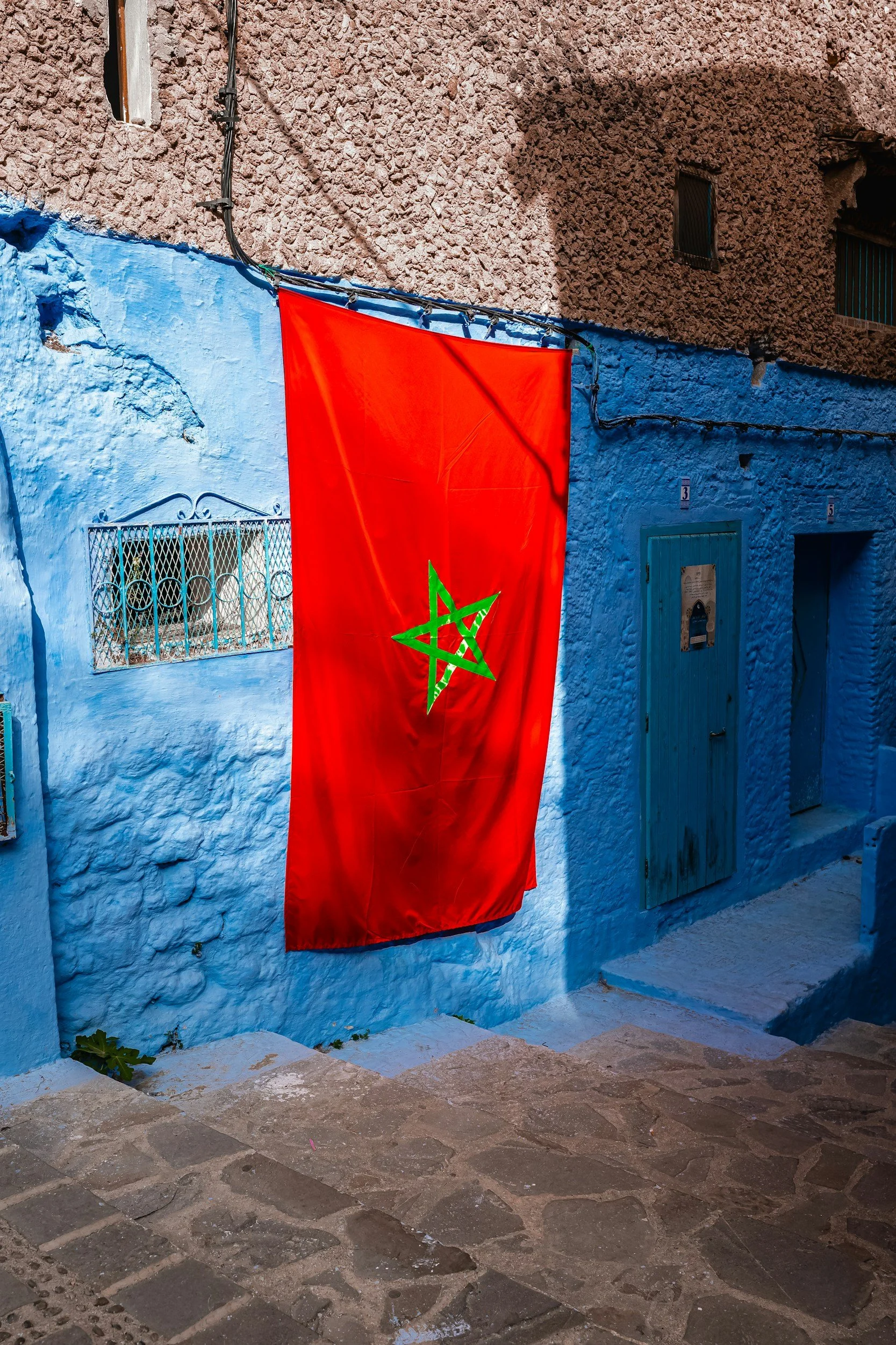 Moroccan flag hanging on a blue wall with cobblestone street in front.