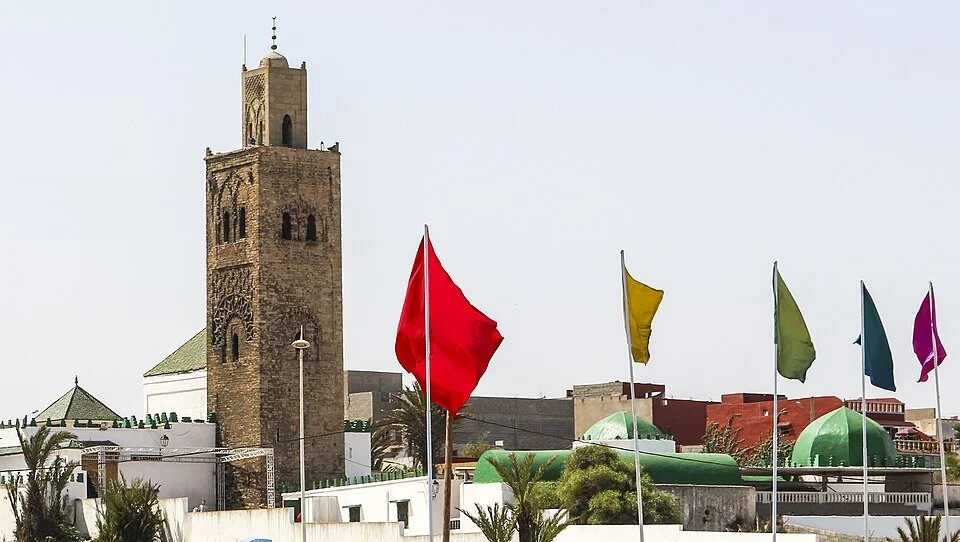 The minaret and green-domed shrine complex of Ribāṭ ʿAyn al-Fiṭr (Tīṭ), Dukkāla coast, with the Moroccan flag in the foreground — the Idrīsī-founded institution where al-Jazūlī spent seven years in formation under Muḥammad Amghār al-Ṣaghīr.