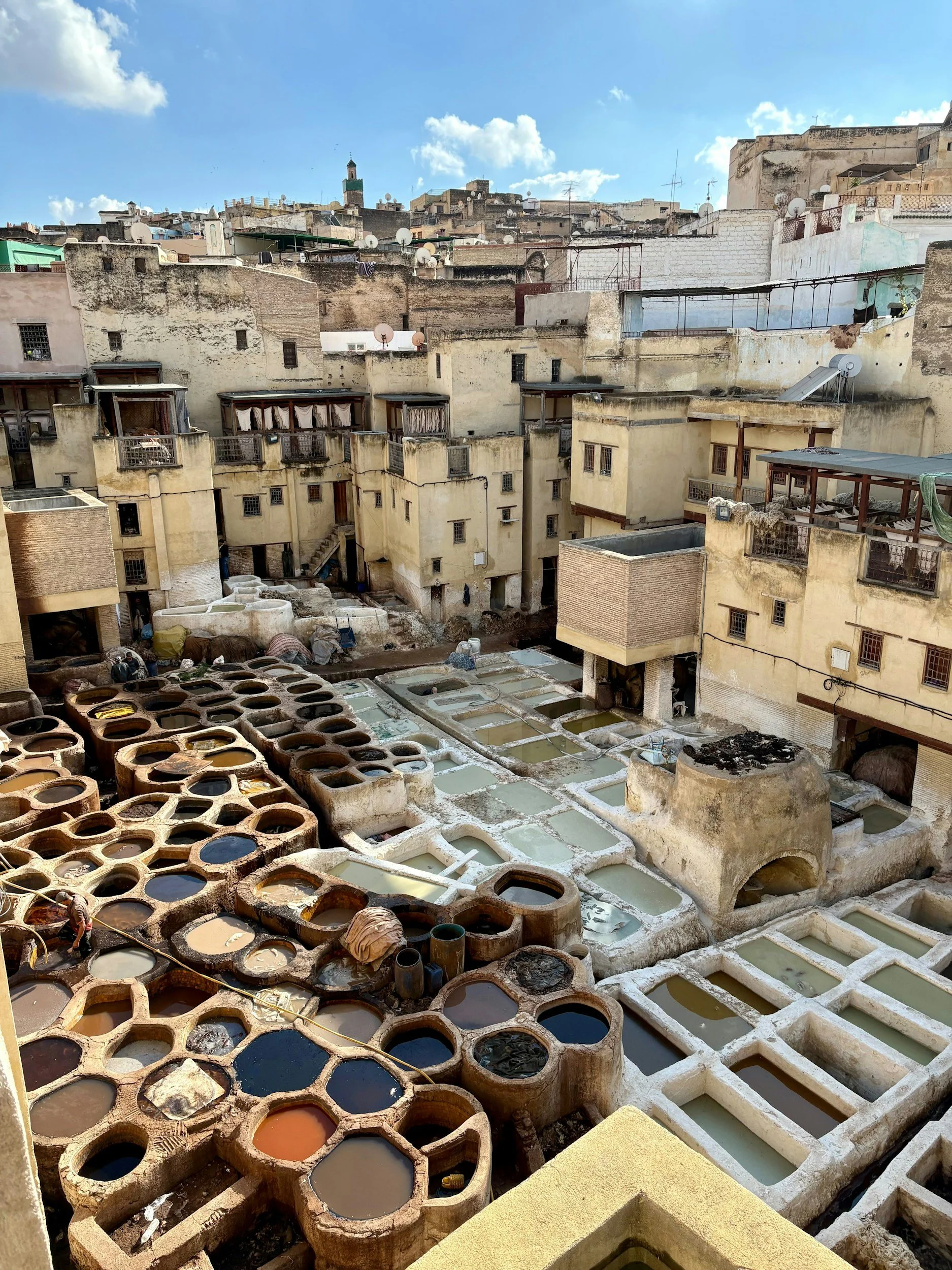 Aerial view of the Chouara tanneries in Fez al-Bālī, Morocco, showing circular stone vats filled with dyes in ochre, white, and blue, surrounded by workers and drying hides.