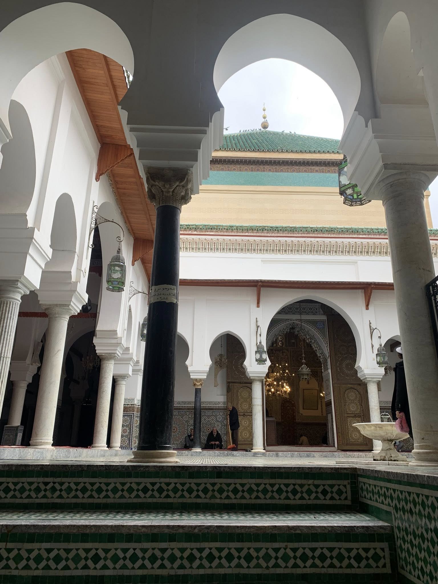 Interior courtyard of the shrine of Mawlāy Idrīs II in Fez al-Bālī, Morocco, showing carved stucco walls, zellige tilework, and a cedarwood screen surrounding the tomb enclosure.