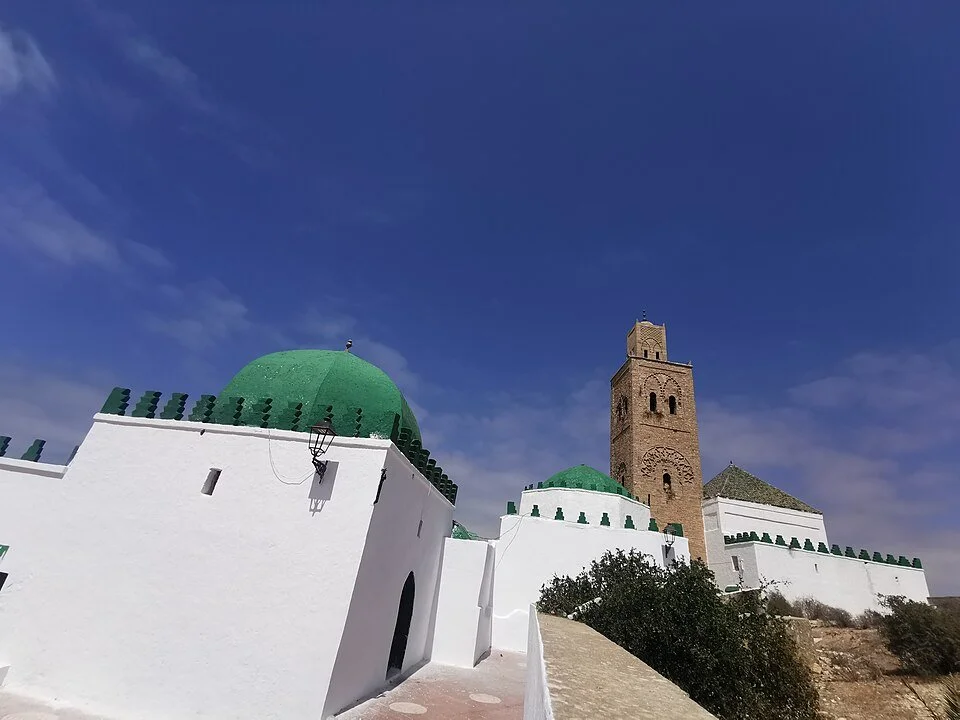 Shrines of the Amghar Ribāṭ (near El Jadida) — A cluster of whitewashed sanctuaries crowned with green domes and edged with green crenellations, set against the Atlantic sky, with a tall stone minaret rising above the complex.