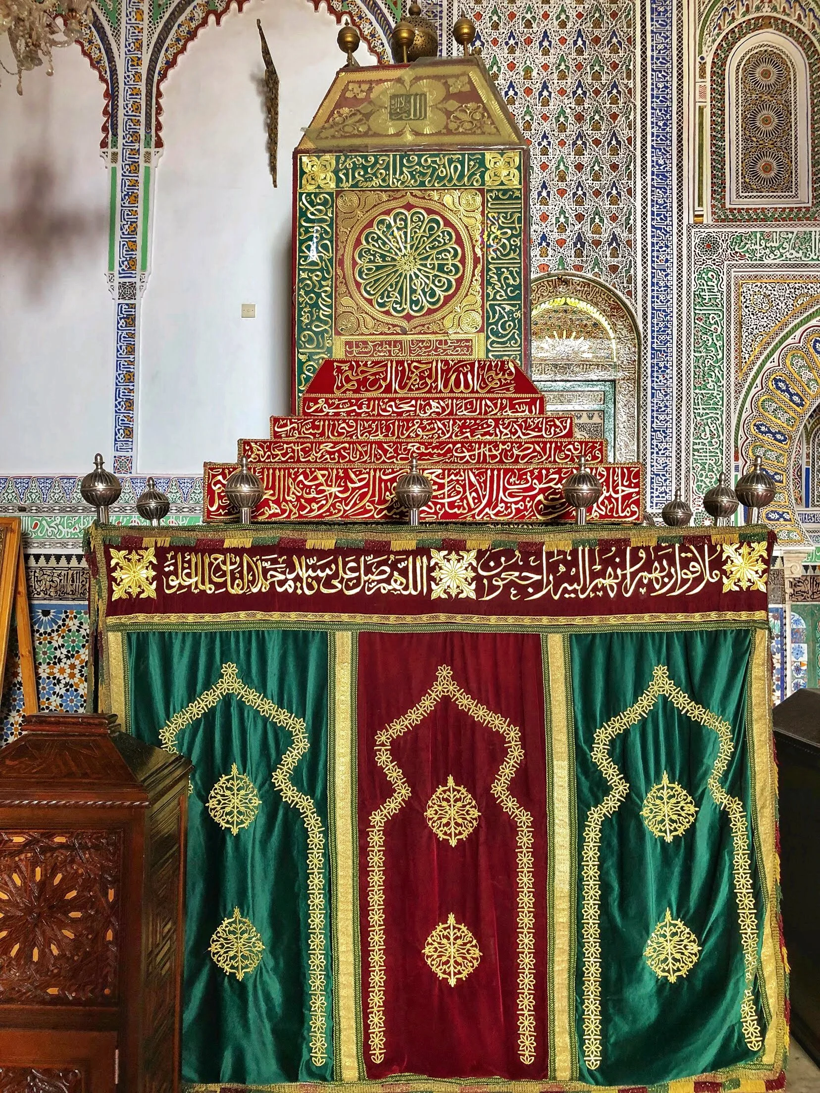 Inside a mosque, a richly decorated mihrab with colorful tiles and a pulpit with embroidered cloth featuring Arabic calligraphy and gold designs.