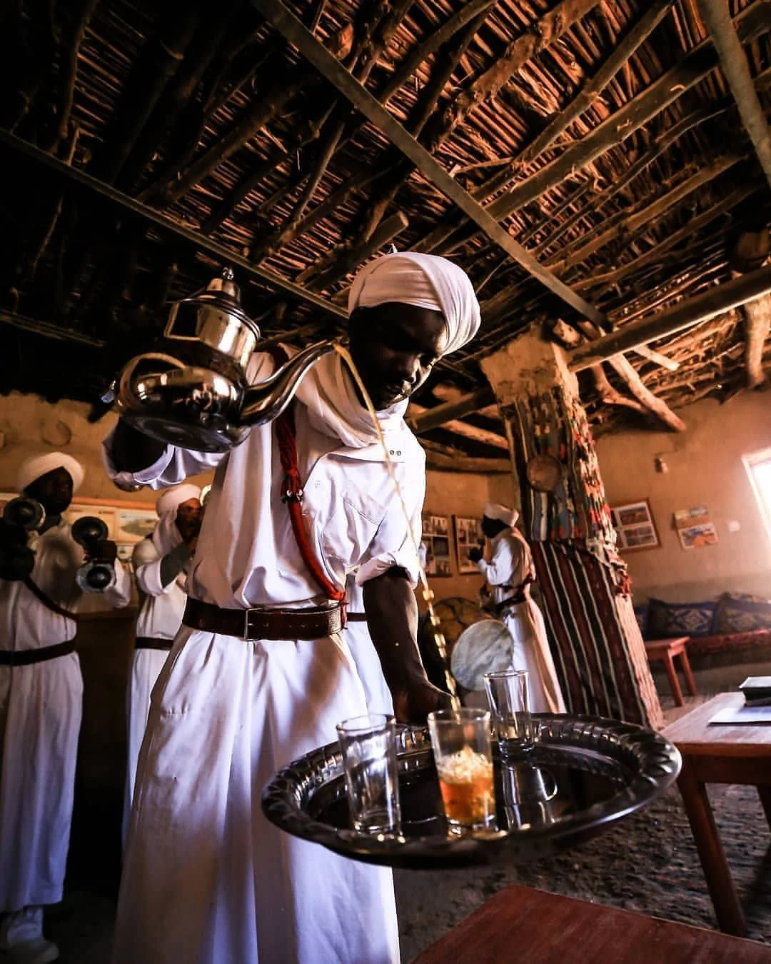 Morocco Tea cutlure: a tayman wearing white traditional clothing and a white turban is pouring tea from a metal teapot into glasses on a tray, inside a rustic room with a wooden ceiling and photographs on the walls.