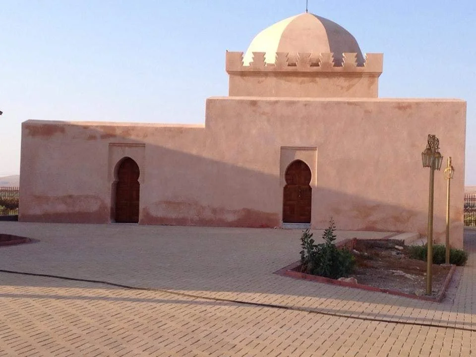 Shrine of Sīdī Shākir — A small maqām built in a plain, earthen style, with thick plastered walls, two horseshoe-arched wooden doors, and a dome rising above a crenellated roofline, opening onto a wide paved courtyard in a calm rural setting.