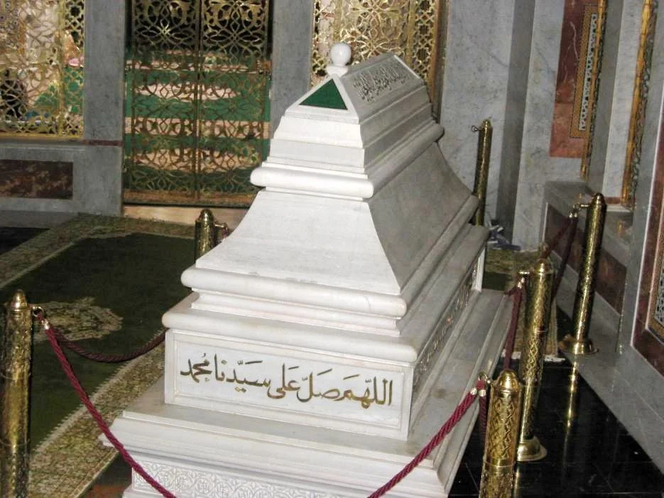 A majestic, tiered white marble tomb of Ahmad al-Tijani, sultan-like in form, enclosed by gold posts and red cords, within an ornate Moroccan shrine rich in geometric tilework and decorative detail.