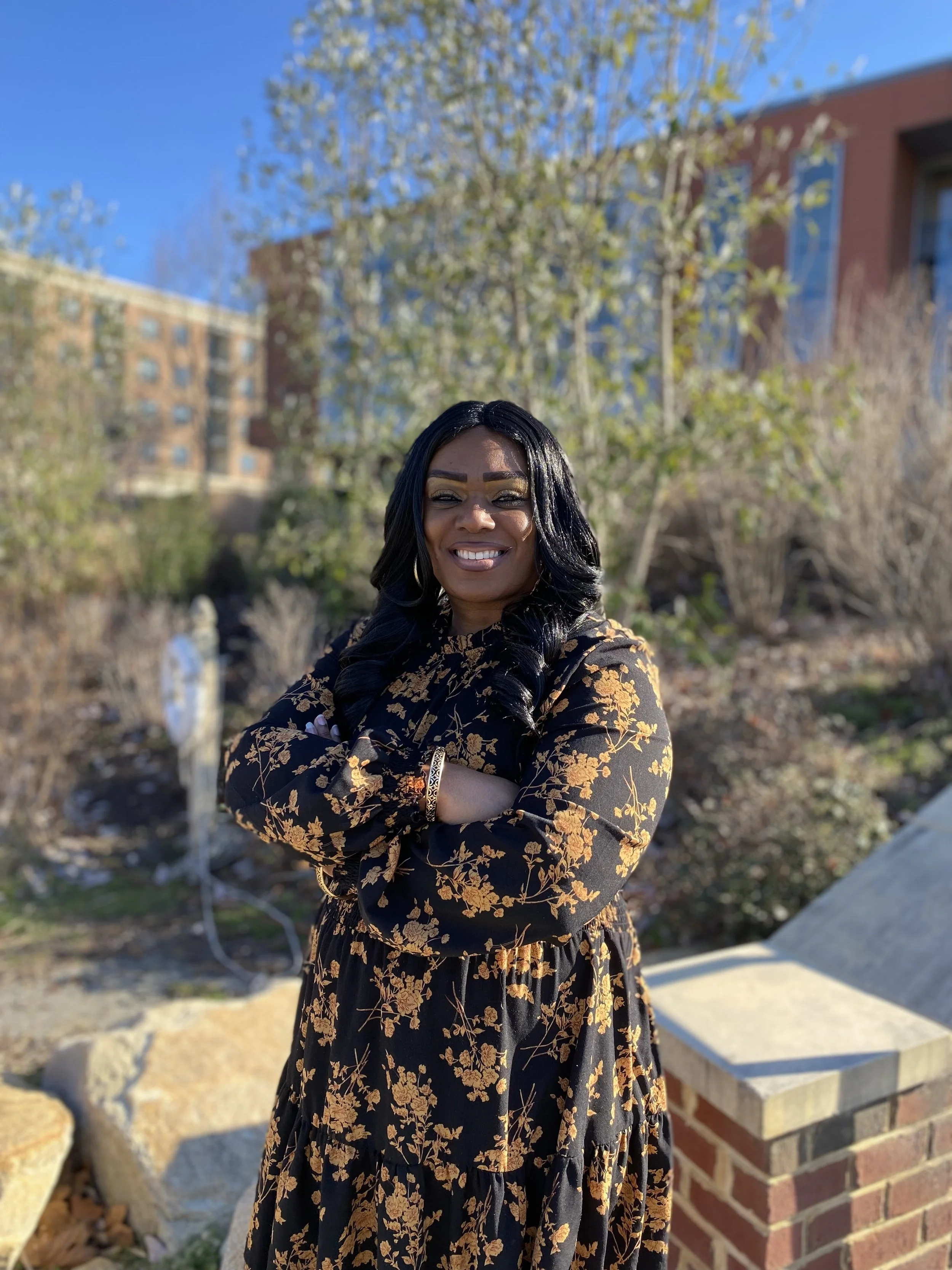A woman with long black hair, smiling and standing outdoors with crossed arms, wearing a black dress with gold floral patterns, in front of trees and buildings on a sunny day.