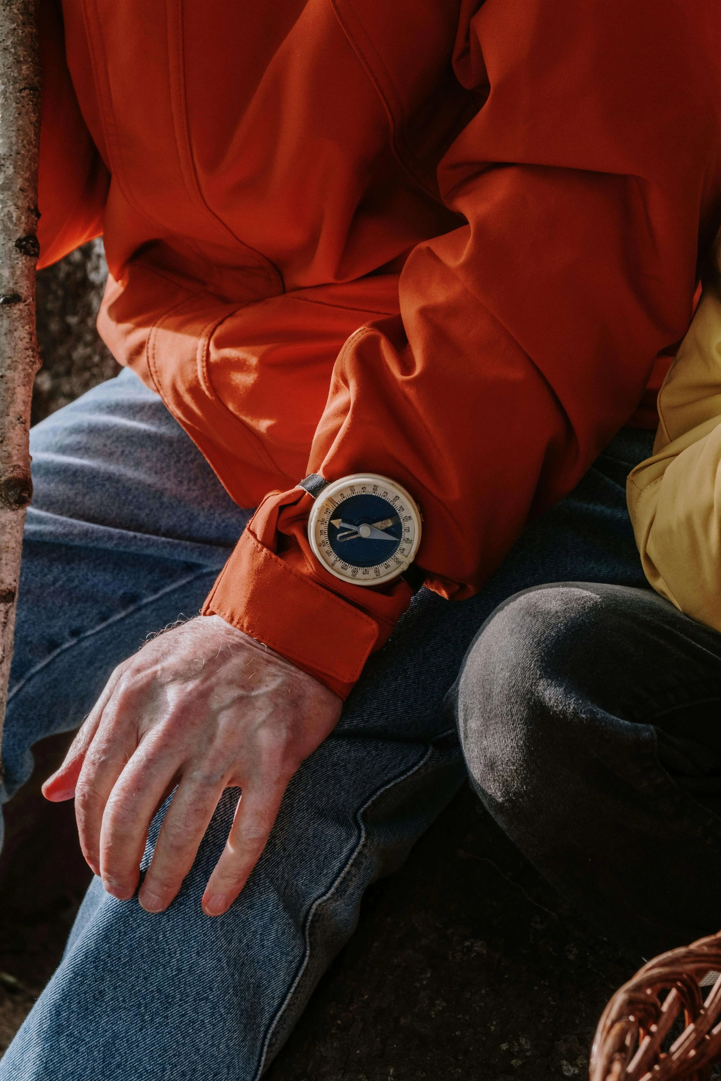 Close-up of a person's arm wearing an orange jacket and a compass on their wrist, with jeans and another person's leg nearby.