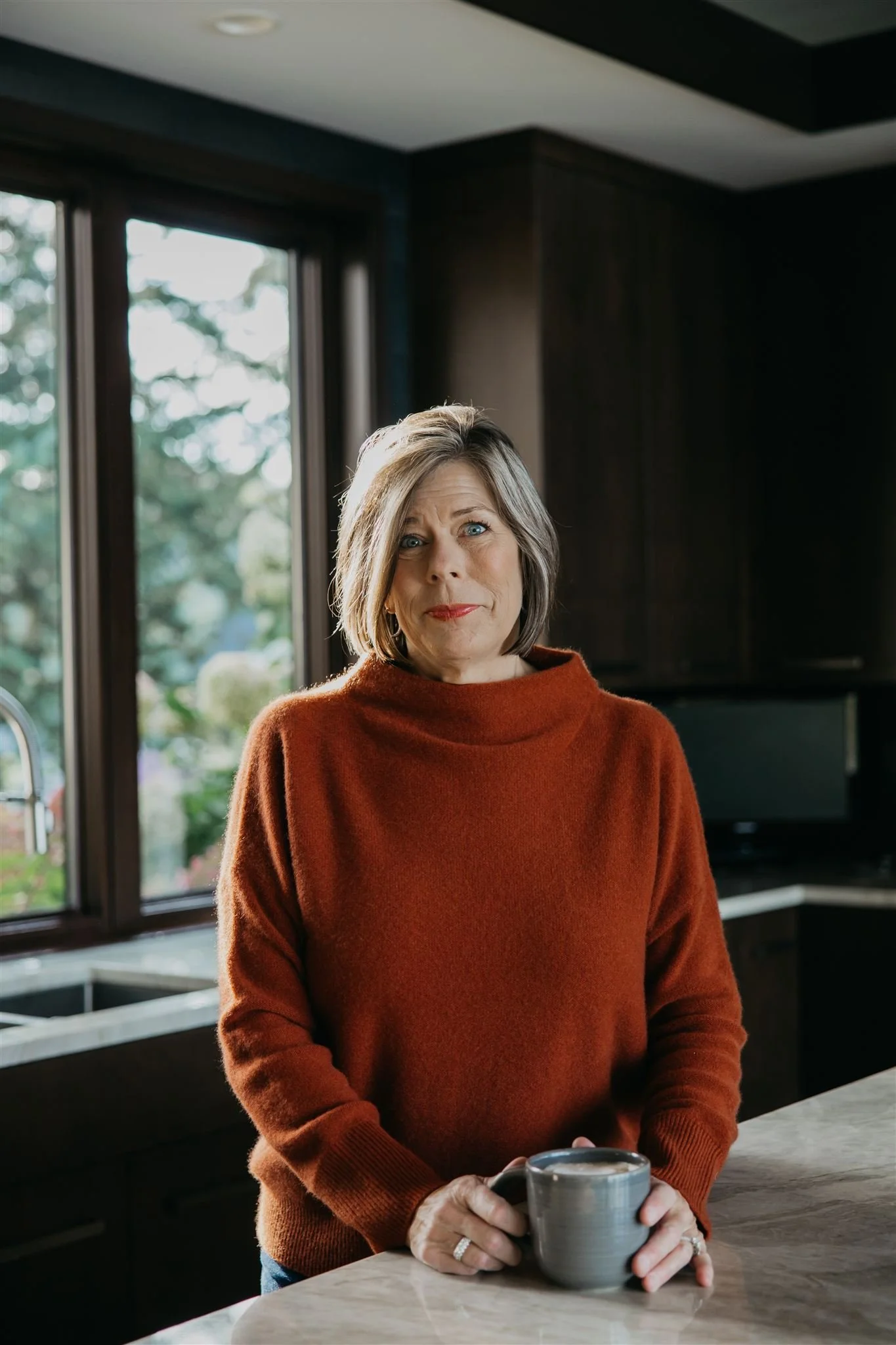A woman with short gray hair and blue eyes standing in a kitchen, holding a gray mug, wearing a rust-colored sweater, with large windows and greenery outside.