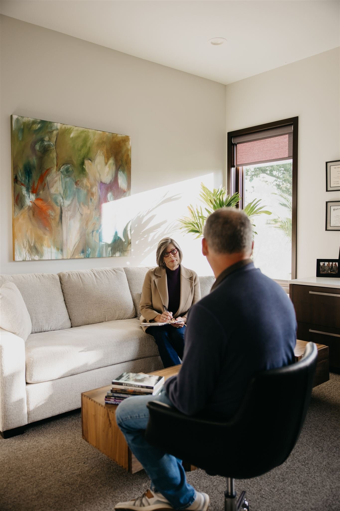 A woman sitting on a beige sofa during a counseling session with a man in a black office chair in a well-lit office with a large window, a colorful abstract painting, and a potted plant.