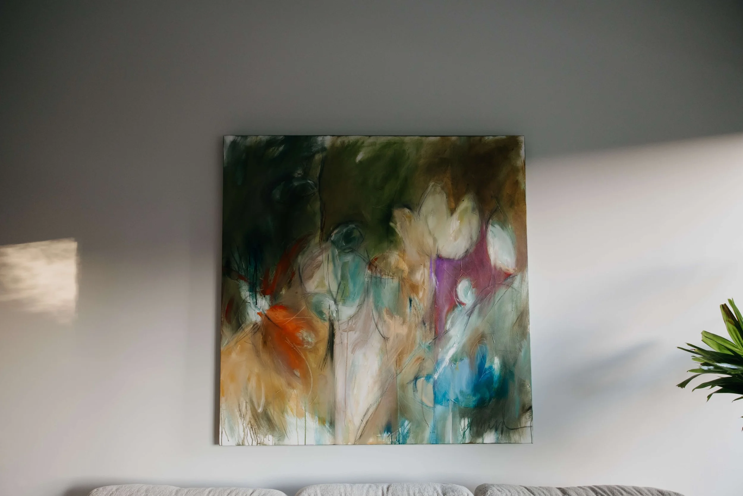 Abstract floral painting hung on a white wall in a living room with a beige couch and a green plant.