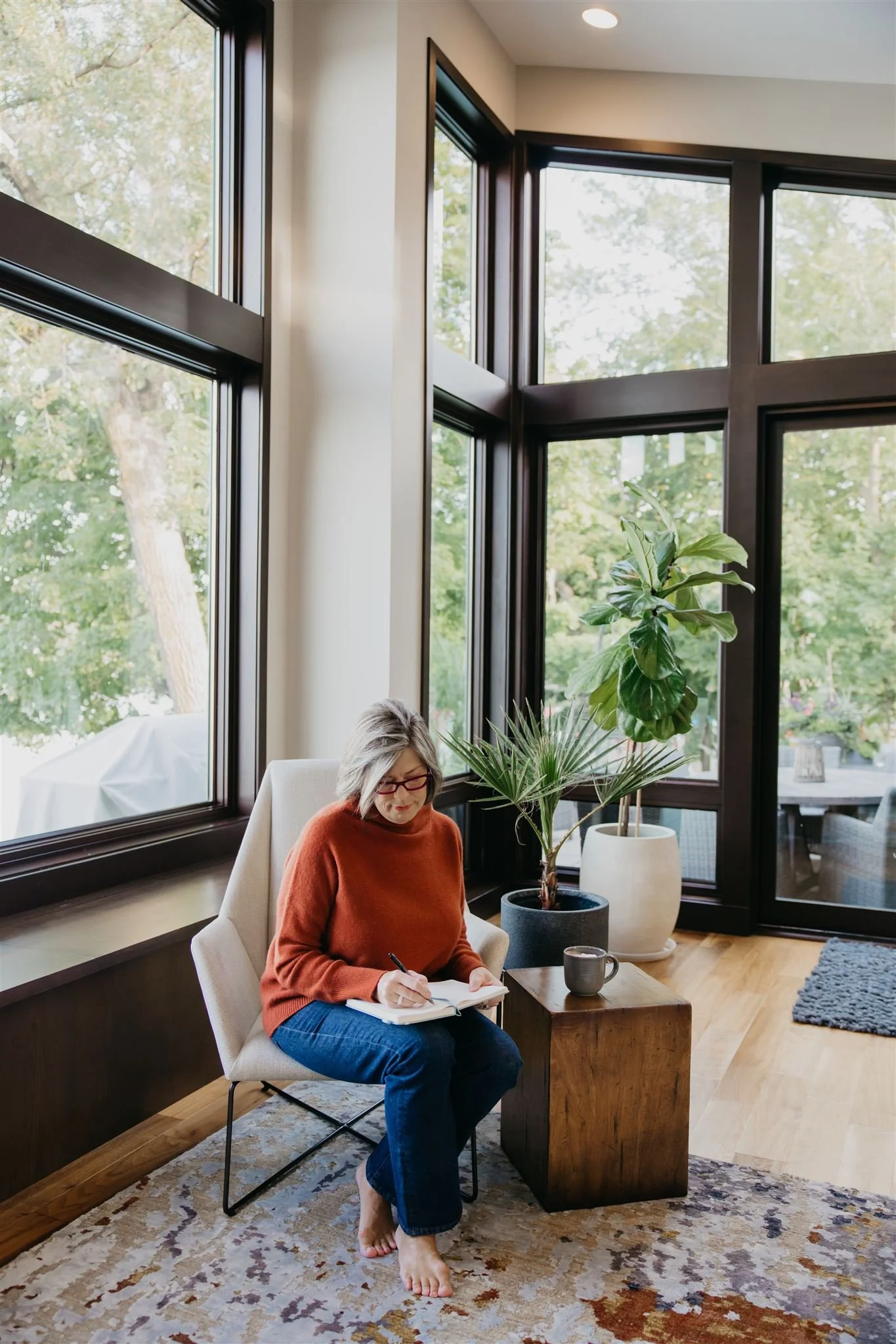 A woman with gray hair, glasses, wearing an orange sweater and blue jeans, sitting barefoot in a beige armchair in front of large windows with greenery outside. She is writing in a notebook with a black pen, and there is a wooden side table with a mug and two potted plants.