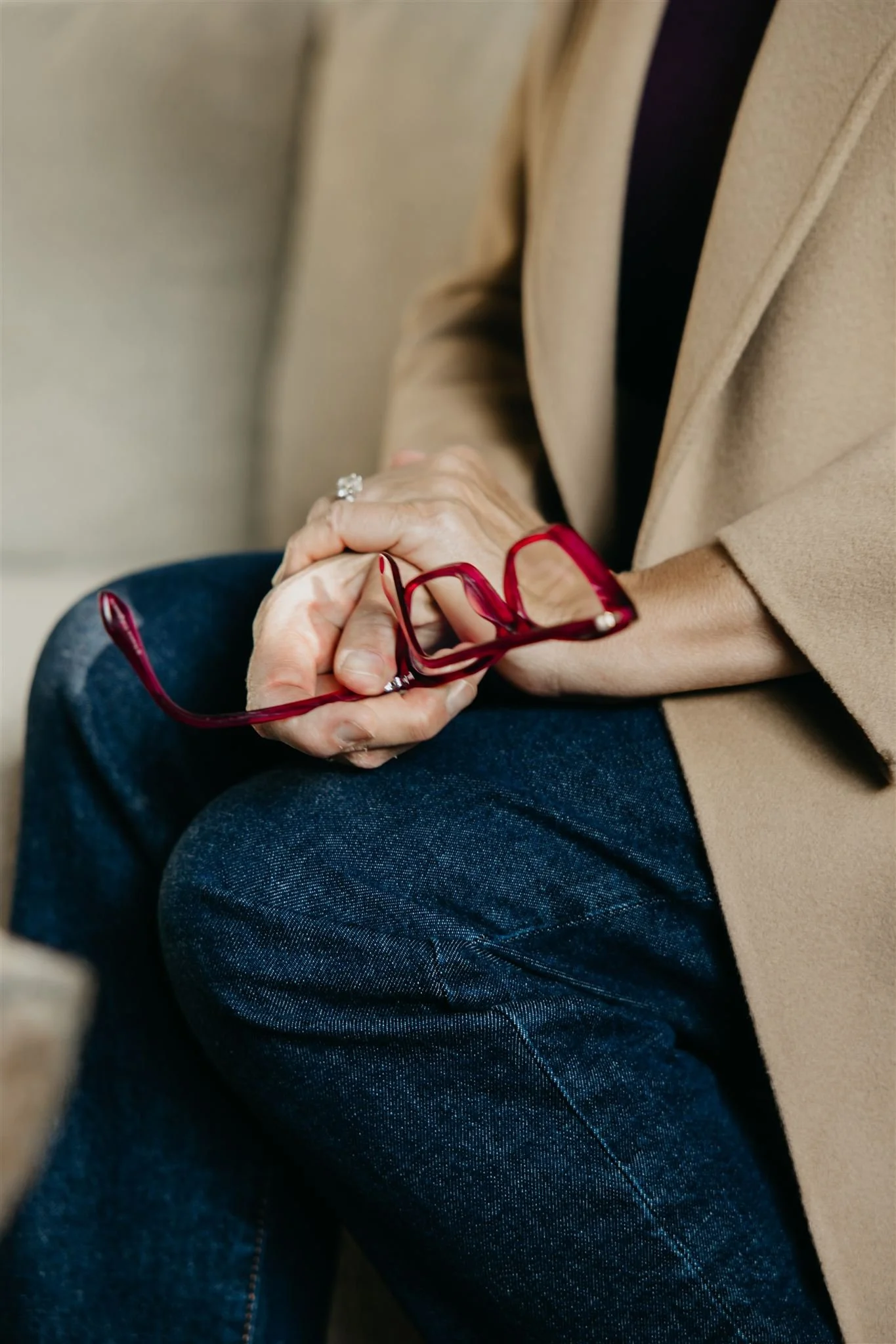 Close-up of a person sitting with hands resting on lap, holding red glasses, wearing a beige coat, dark shirt, and blue jeans.