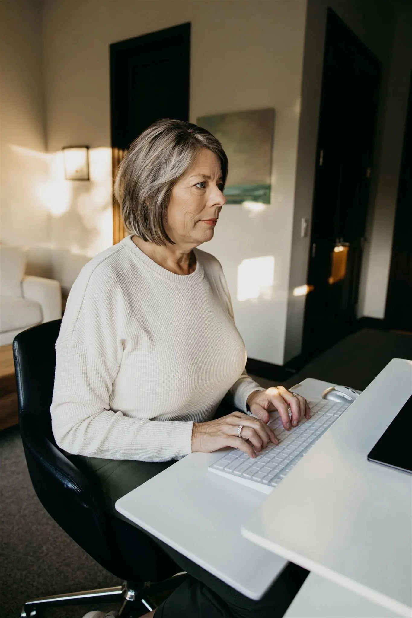 An older woman with shoulder-length gray hair working on a white computer keyboard at a desk in a room with beige walls and artwork.