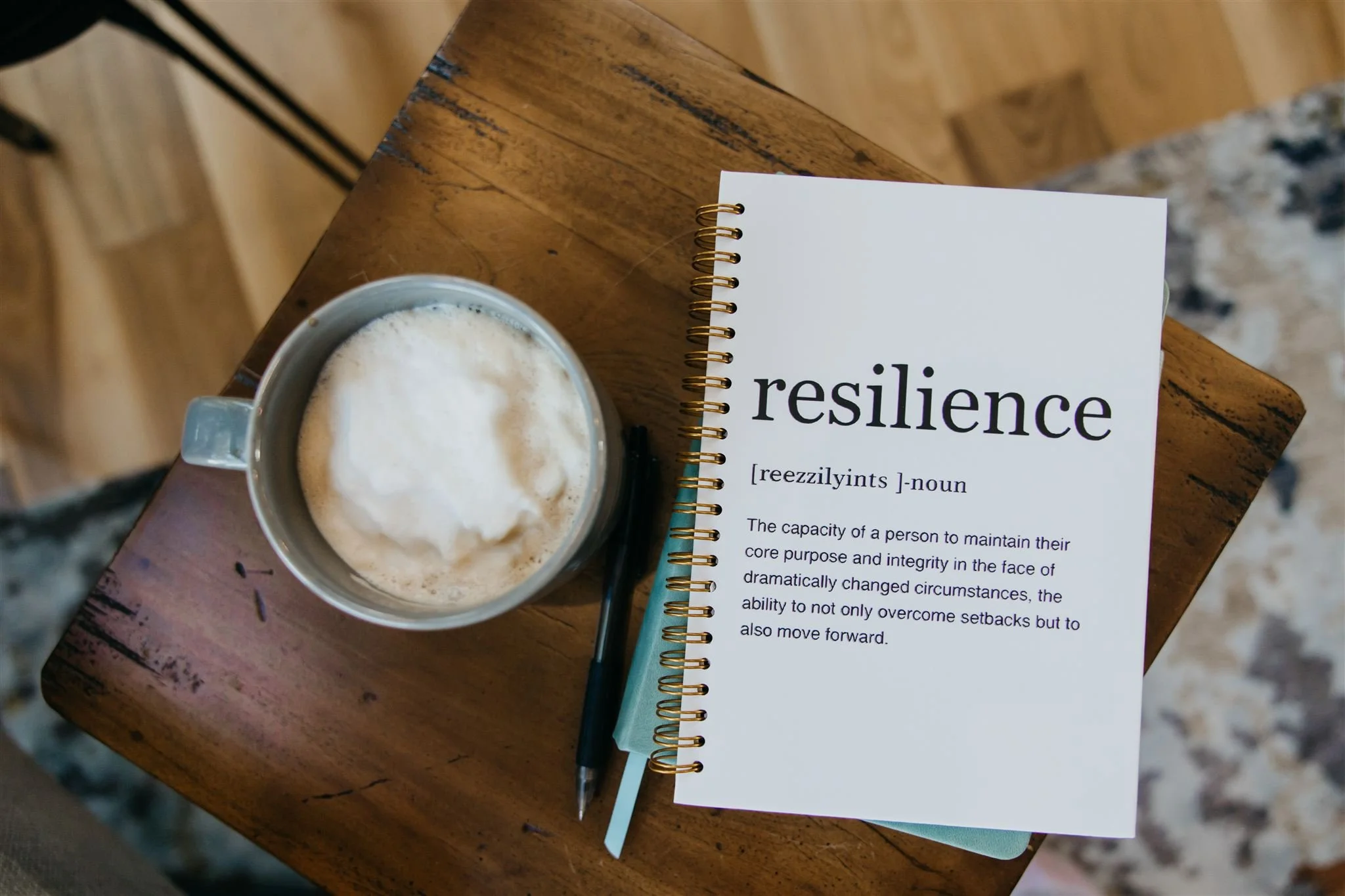 A wooden table with a mug of frothy coffee, a notebook titled 'resilience' with its definition, and a pen beside the notebook.