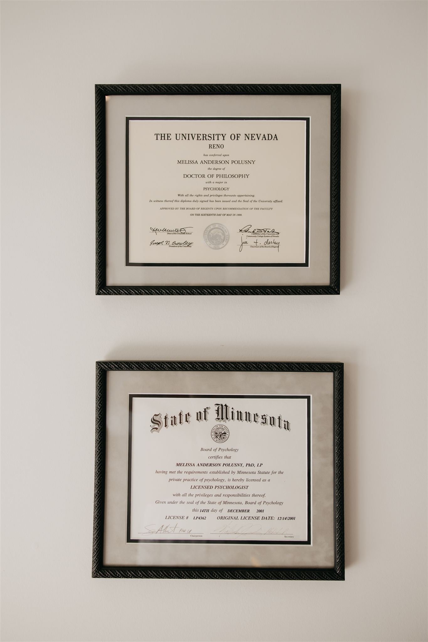 Two framed certificates on a white wall. The top certificate is from the University of Nevada, Reno, awarded to Melissa Anderson Polusny for a PhD in psychology. The bottom certificate is from the State of Minnesota, certifying Melissa Anderson Polusny as a licensed psychologist, dated December 14, 2001.