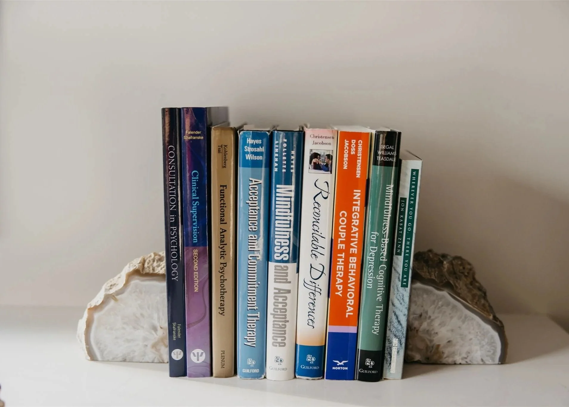 A row of psychology and therapy books on a white shelf, supported by two decorative white mineral rocks, against an off-white wall.