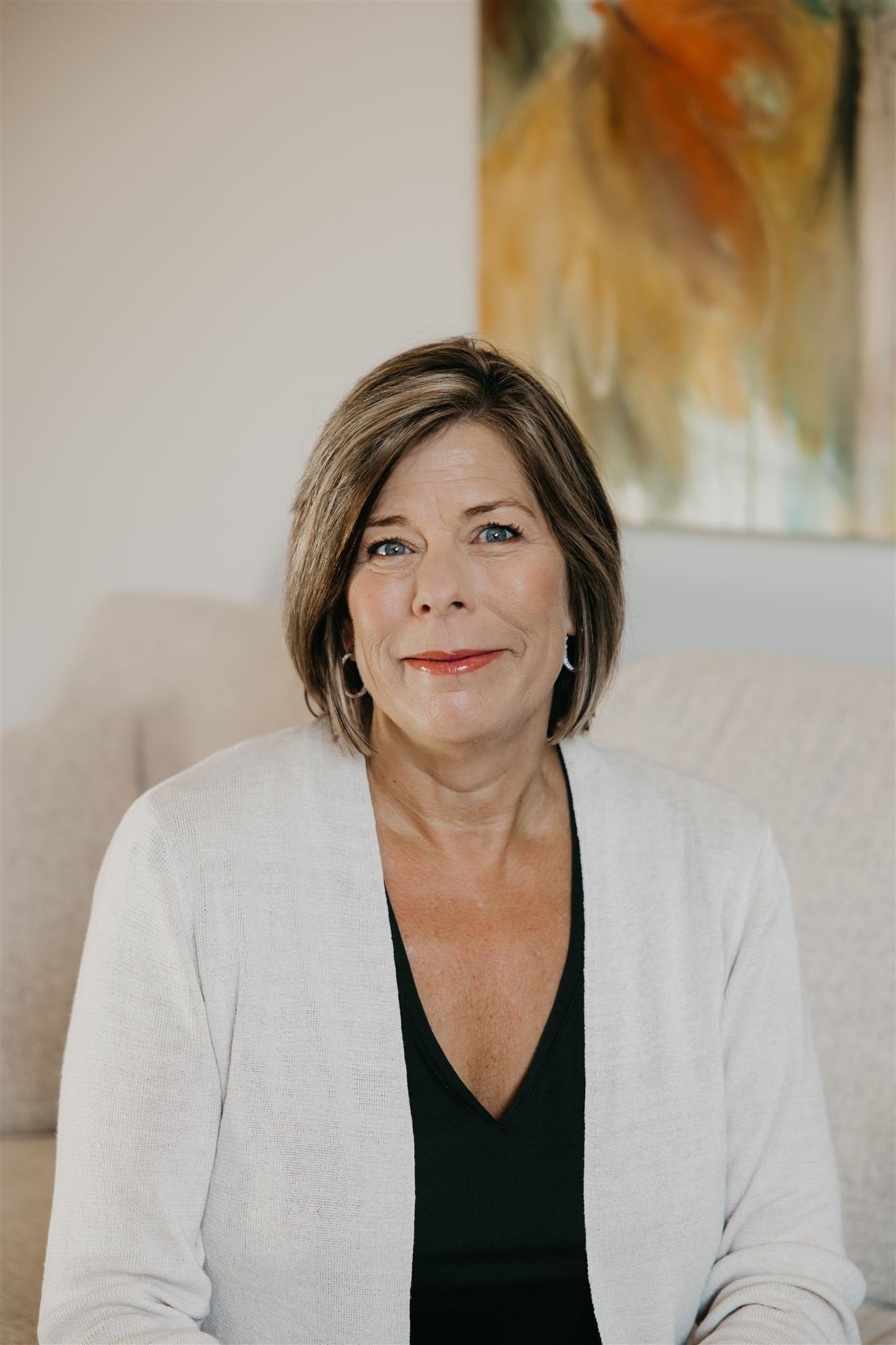 A middle-aged woman with short brown hair and blue eyes sitting on a beige sofa, smiling, with a colorful abstract painting on the wall behind her.