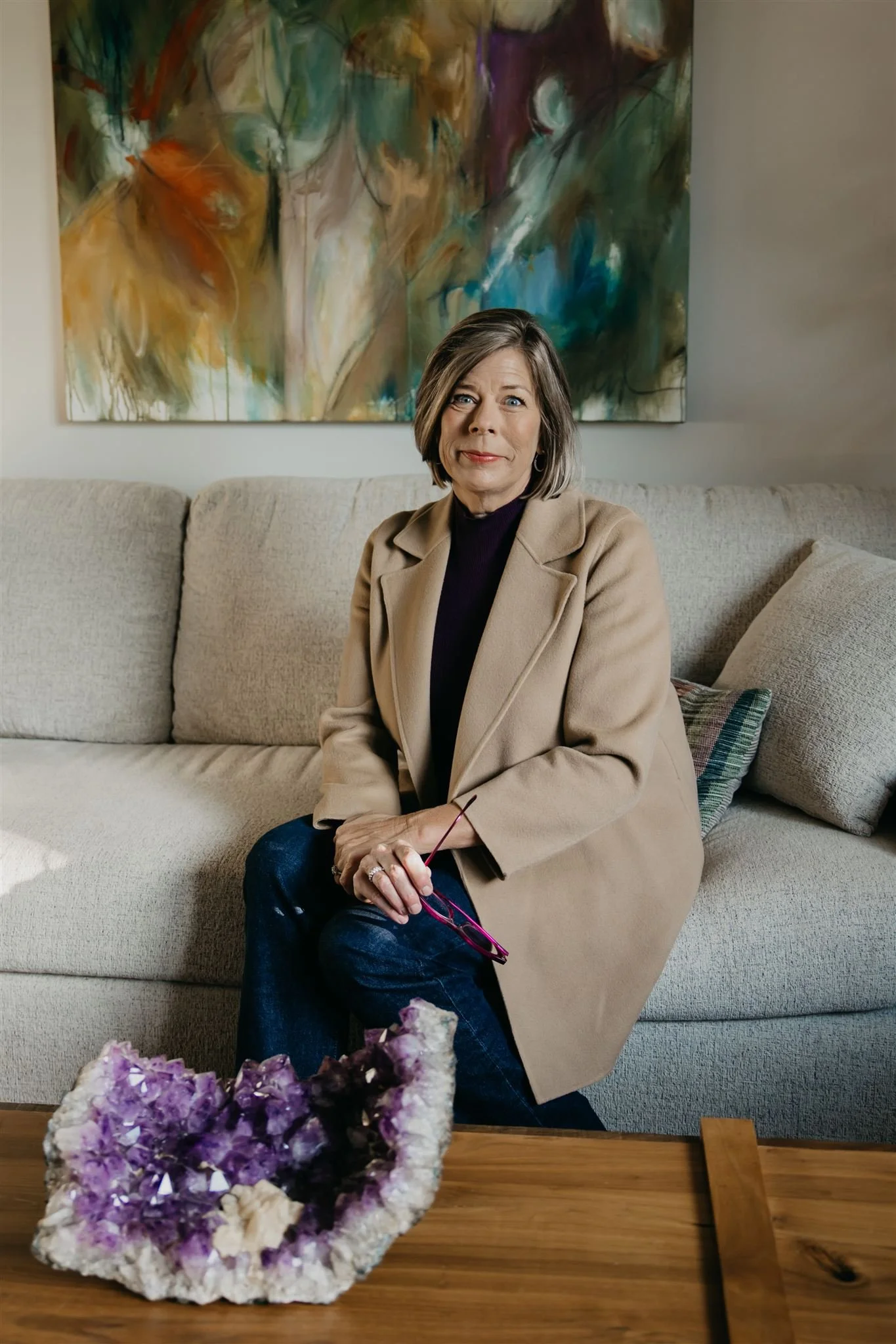 A woman sitting on a beige sofa in a living room, holding pink glasses, with colorful abstract artwork behind her and a purple amethyst geode on a wooden table in front of her.