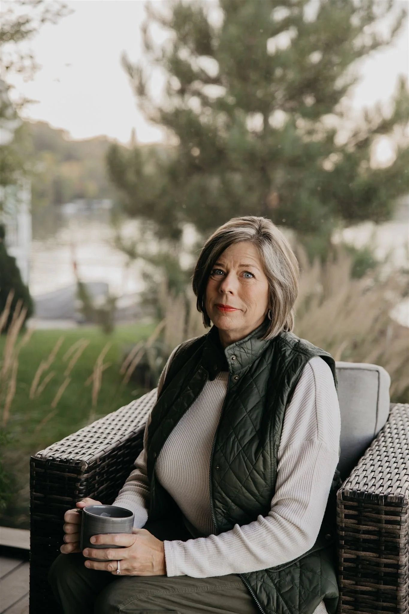 A woman sitting on a wicker chair outdoors, holding a coffee mug and looking at the camera with a slight smile.