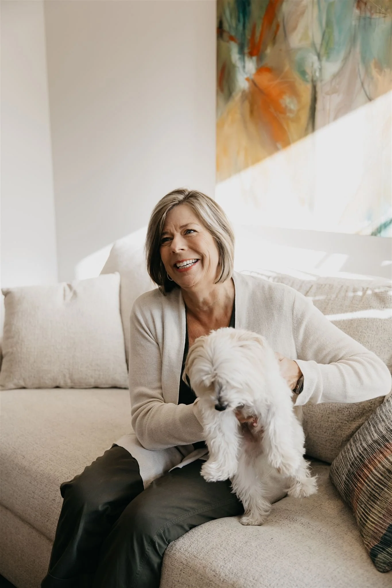 A smiling woman holding a small white dog on a beige sofa in a well-lit living room.
