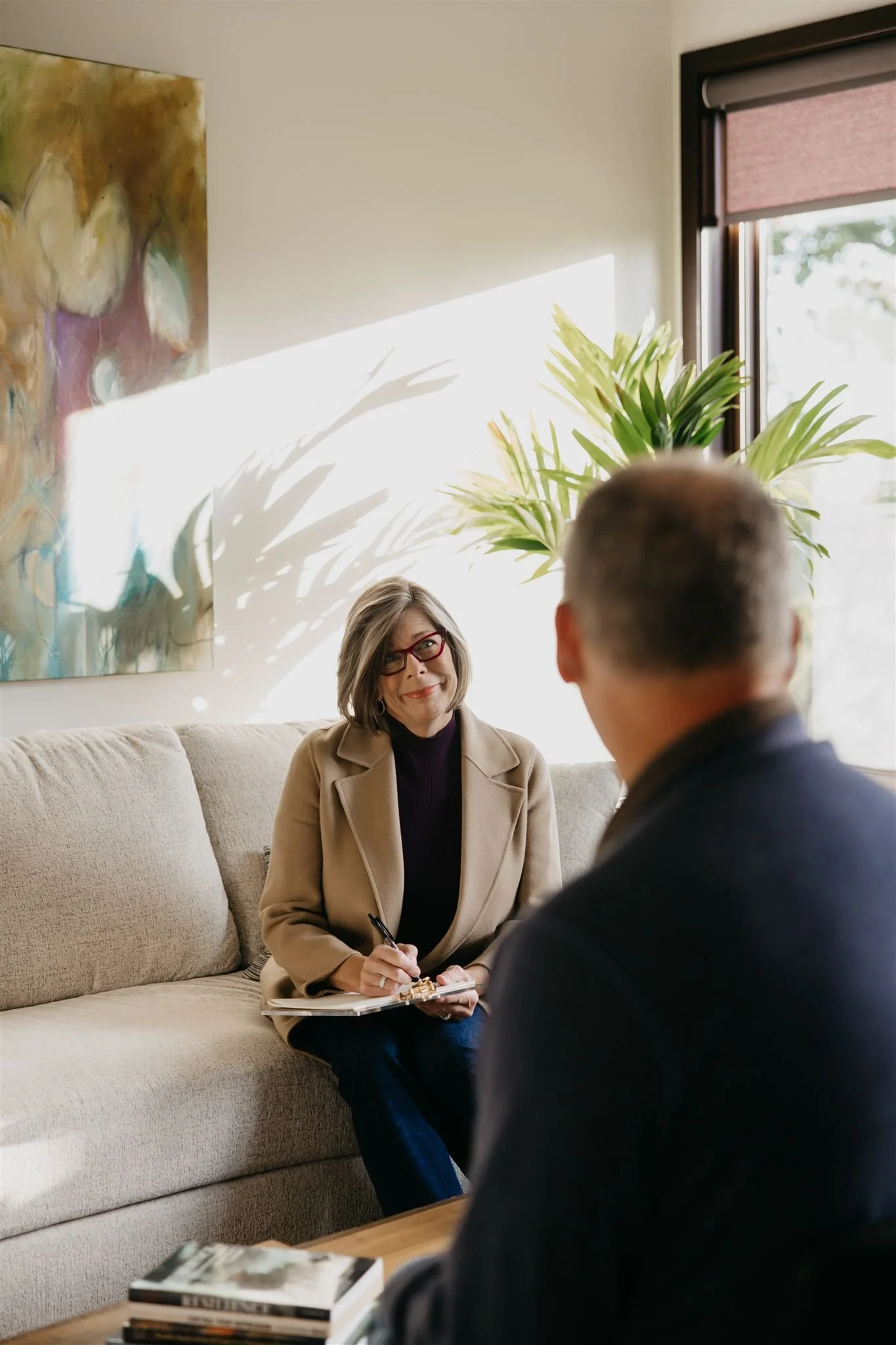 A woman with glasses and a beige blazer smiling during a conversation with a man in a dark jacket in a cozy, well-lit living room.