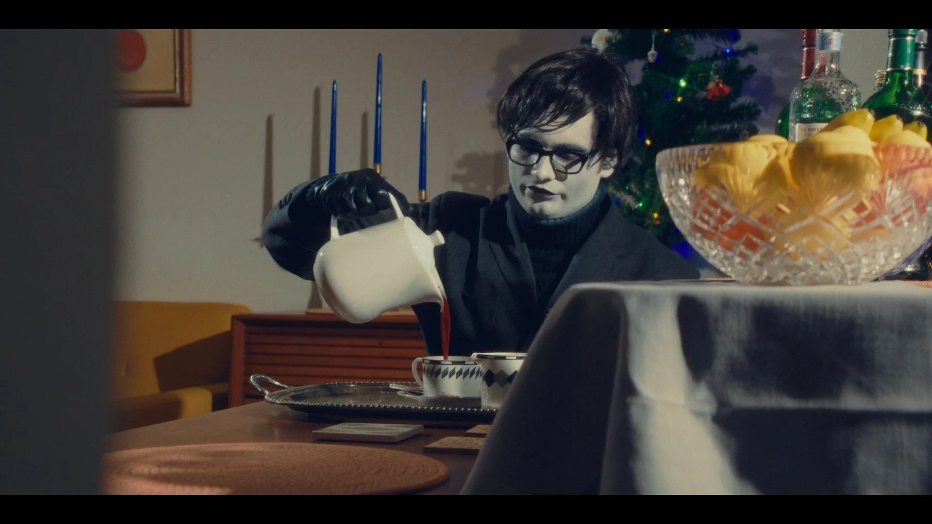 A person with black hair and glasses pouring red liquid into a cup at a festive dining table, with a Christmas tree and decorations in the background.