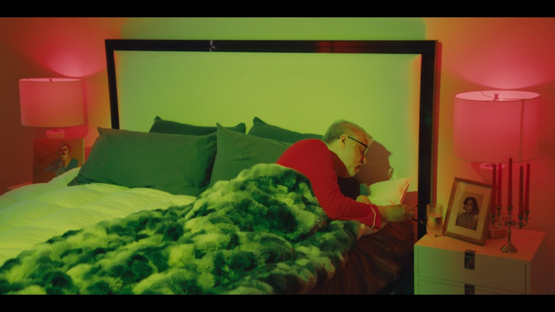 An elderly man with glasses and a gray beard is lying on his stomach on a bed, looking at a tablet. The bed has green pillows and a plush green blanket. There are two pink lamps on nightstands on either side of the bed, with one nightstand also holdi