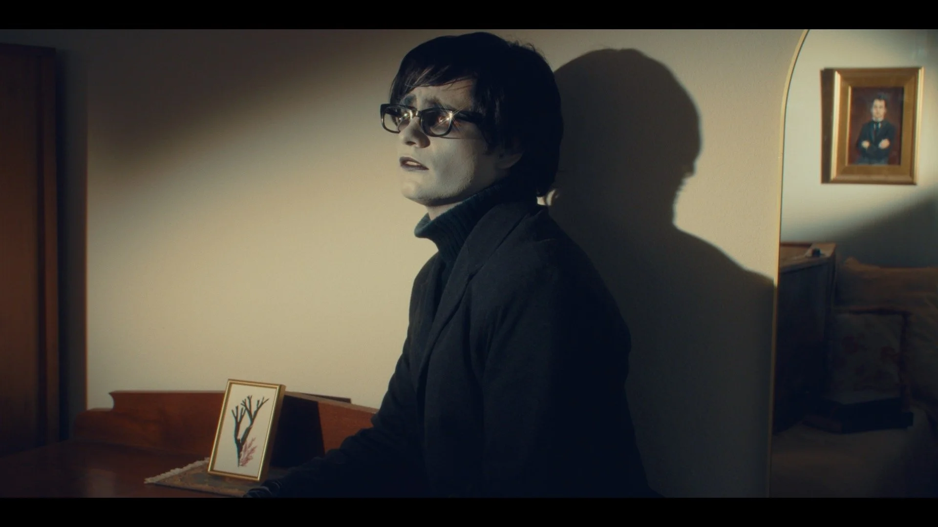 A man with black hair and glasses, dressed in black, sits against a wall with a serious expression, casting a shadow on the wall. There are framed pictures and a small painting on the table and wall behind him.