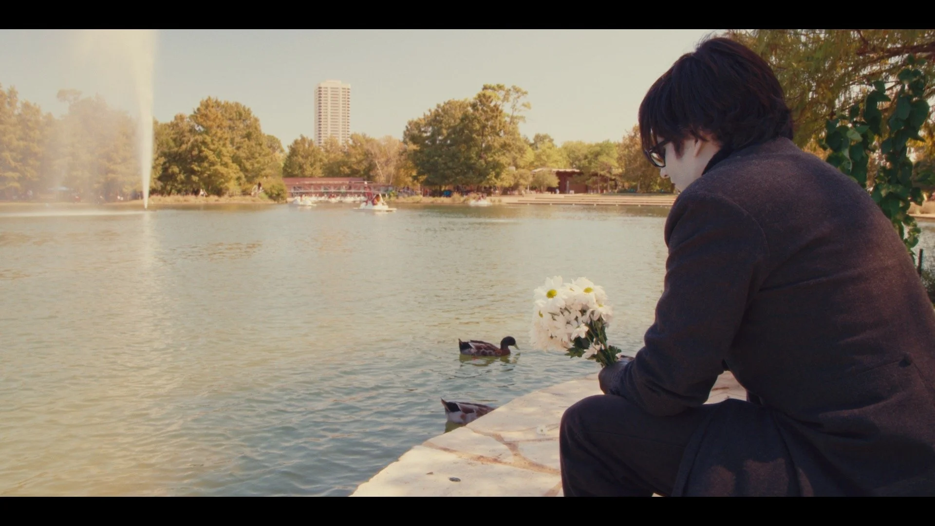 A person with dark hair wearing glasses and a dark jacket holds a bouquet of white daisies while sitting by a lake, watching ducks swim.