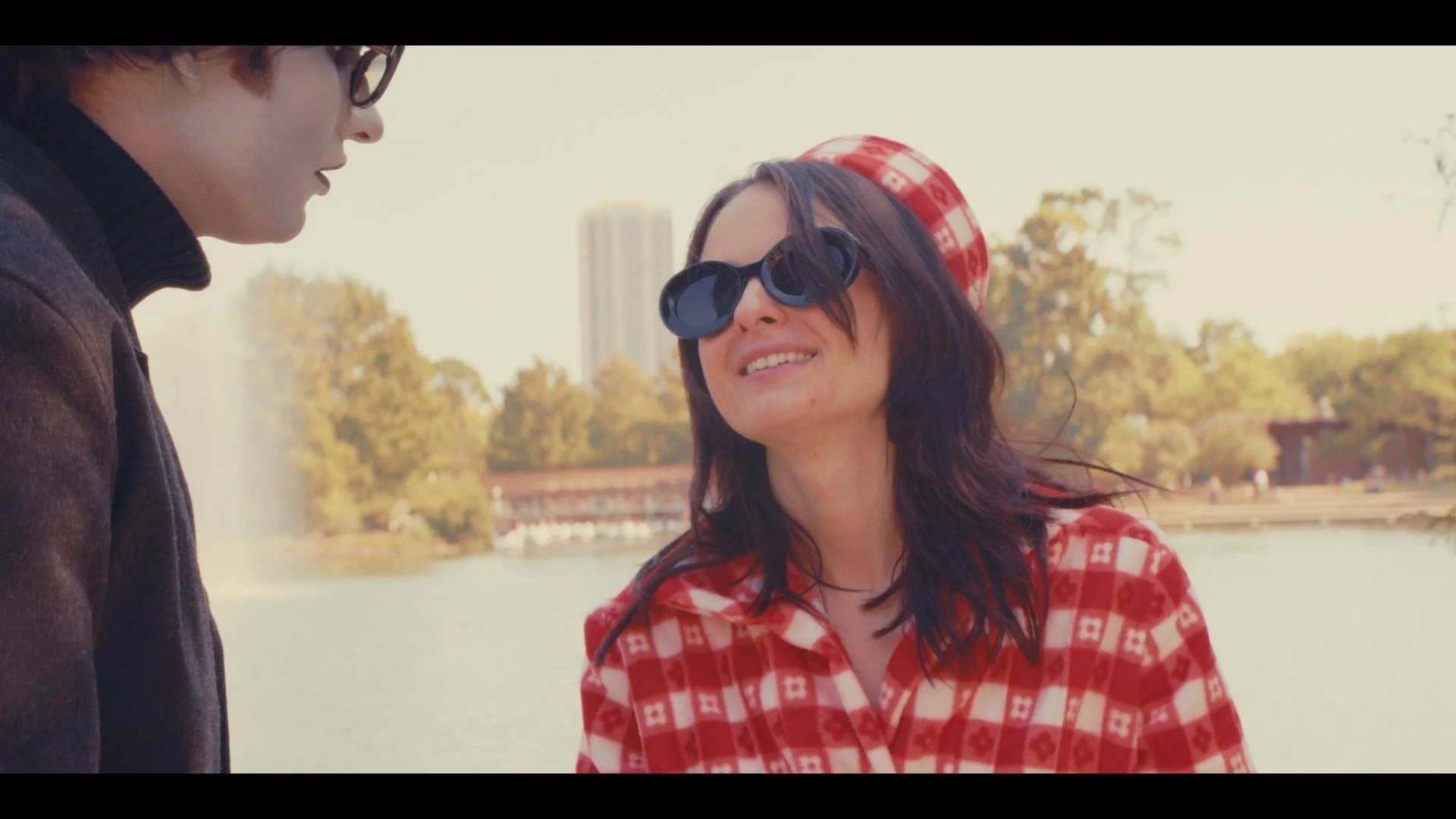A woman wearing large sunglasses and a red checkered jacket smiling and looking at a man near a lake with trees and buildings in the background.
