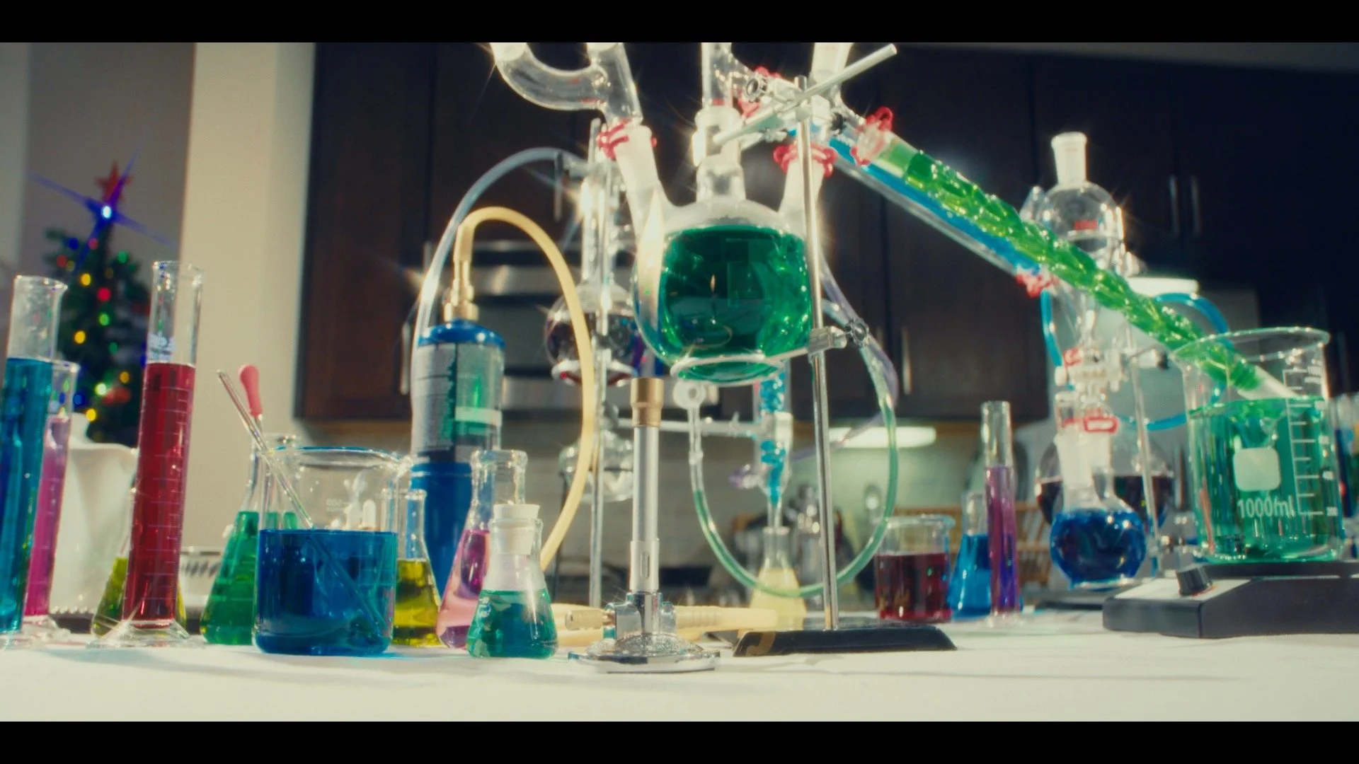 A colorful chemistry laboratory setup with various glassware containing different colored liquids, and a Christmas tree in the background.