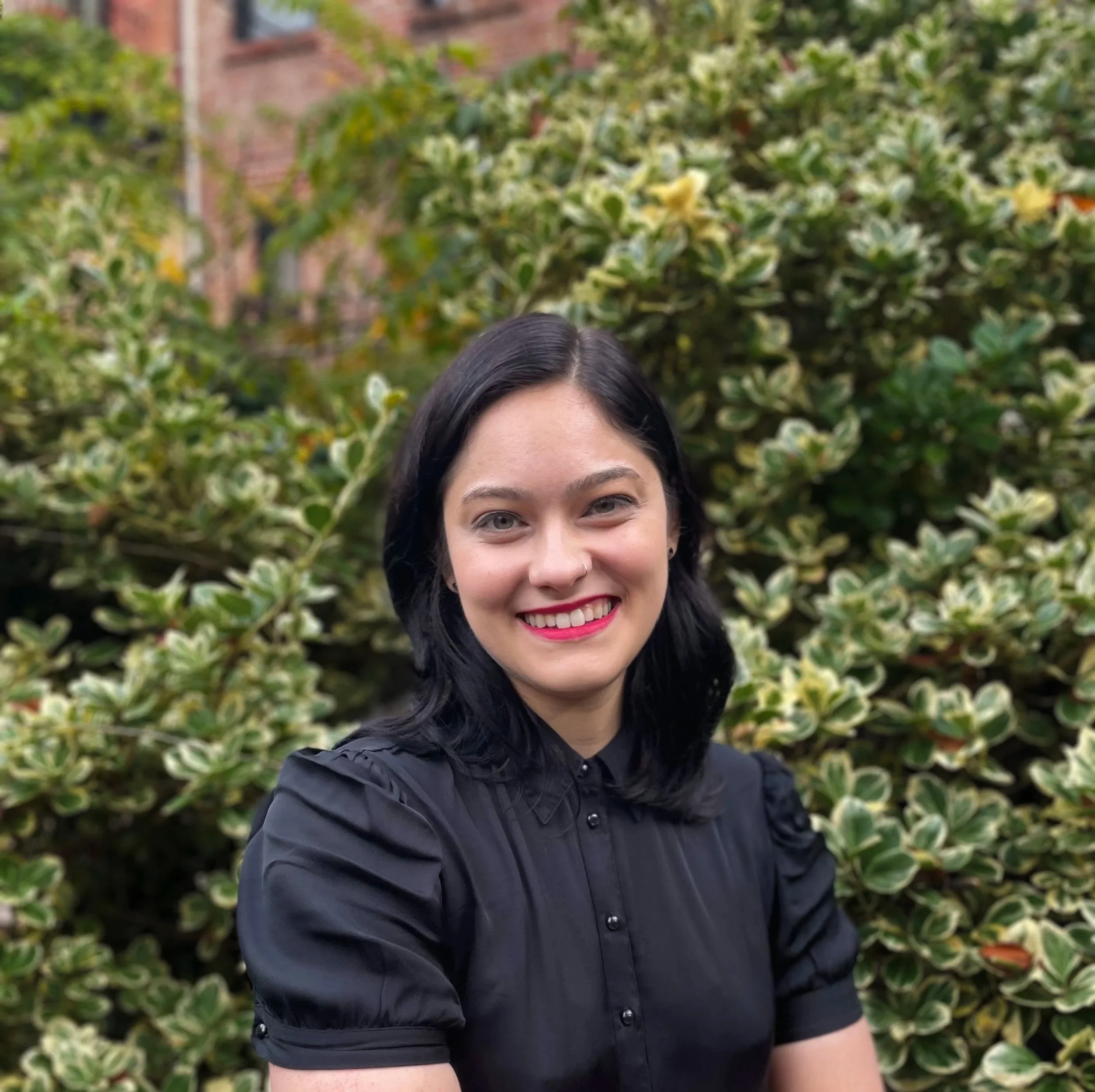 Close-up of ADHD coach Alexandra Schoenborn of Joriki Coaching taken in Brooklyn, NY. She has dark hair and light eyes and is smiling outdoors with green leafy bushes in the background.