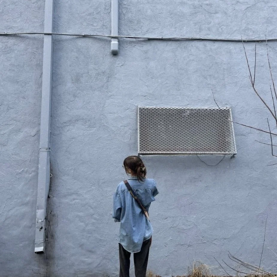 A woman in a denim shirt and dark pants standing with her back to the camera, facing a gray concrete wall with a vent and some branches on the right side.