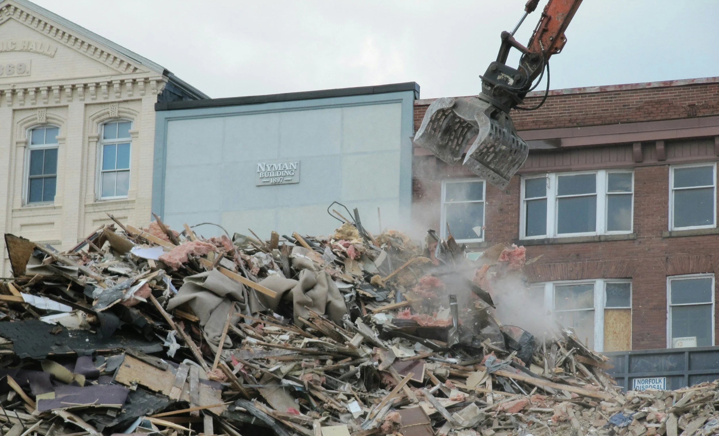 Construction debris and rubble being cleared by heavy machinery outside a building with a sign reading 'Nyman Building 1897'.