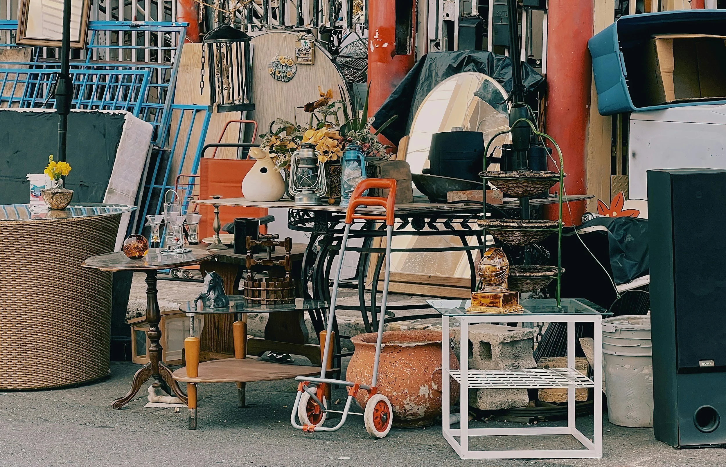 An outdoor flea market or thrift store display with various furniture, decorative items, and household objects arranged on the ground and stacked against a wall.